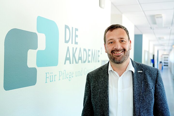 Porträt von Andreas Heck, Geschäftsführer der AKADEMIE² für Pflege und Gesundheit. Er steht lächelnd im Flur der Pflegeschule, an der Wand ist das Logo der AKADEMIE² für Pflege und Gesundheit zu sehen.