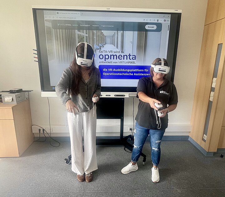 Zwei Frauen mit aufgesetzten VR-Brillen stehen im Unterrichtsraum vor einem Bildschirm mit der Browseransicht von opmenta. In den Händen halten sie Controller, mit denen sie das Anordnen von Instrumenten auf dem virtuellen Instrumententisch simulieren.
