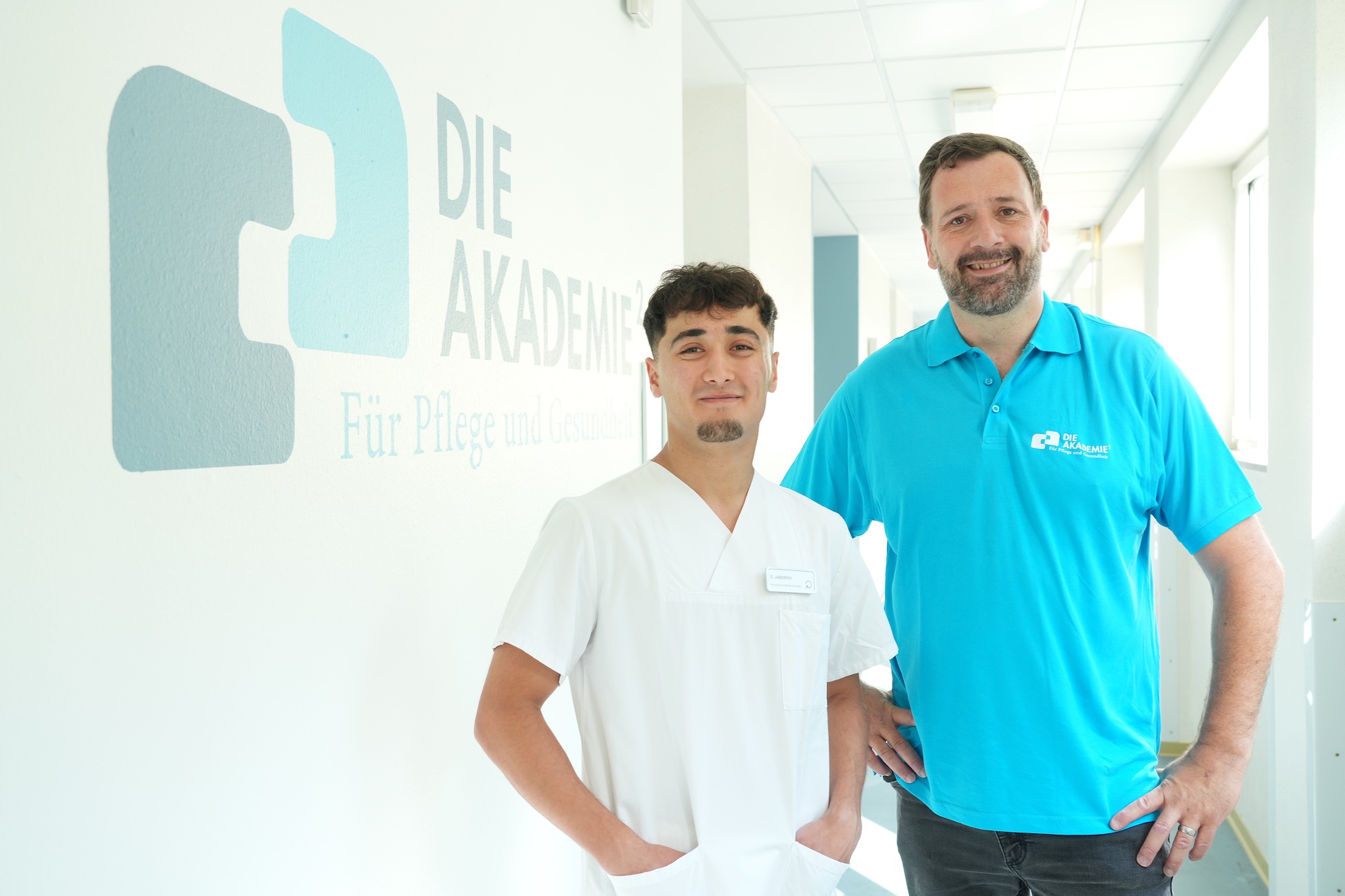 Zwei Männer stehen nebeneinander im Flur der AKADEMIE². Der jüngere trägt einen weißen Kasack, der ältere ein türkisfarbenes T-Shirt.