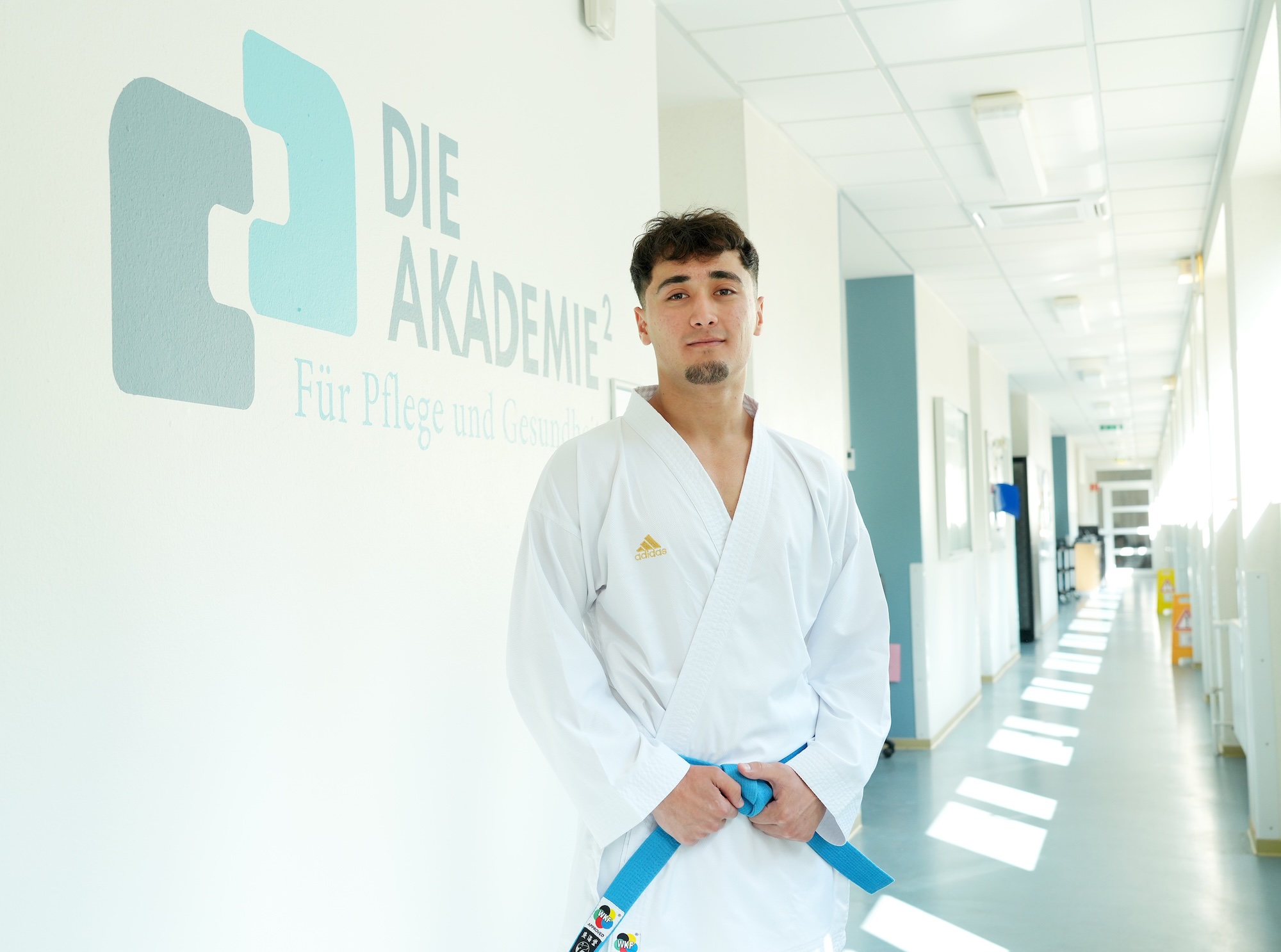 Ein junger Karateka steht auf einem Flur vor einer Wand mit dem Logo der AKADEMIE². Er hält seinen Karategürtel mit beiden Händen.
